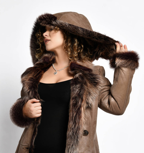 Fashionable woman with curly red hair in bodysuit poses holding pulling a fur hood of her sheepskin coat with fur cuffs - Foto, afbeelding