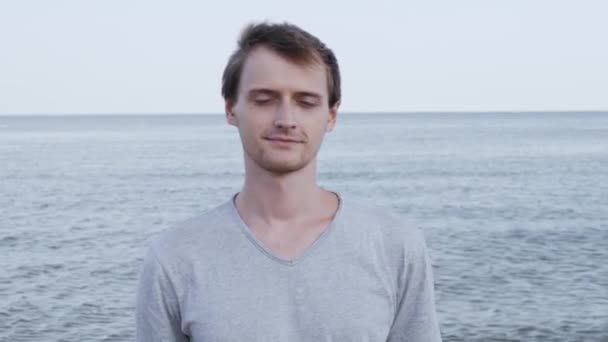 Πορτρέτο του ελκυστικός γοητευτικός νέος Καυκάσιος άνθρωπος χαμογελαστός στην ήρεμη παραθαλάσσια παραλία στο ηλιοβασίλεμα φορώντας γκρι t-shirt - Πλάνα, βίντεο