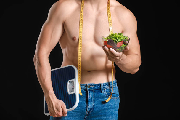 Handsome muscular man with measuring tape, salad and scales on dark background. Weight loss concept - Fotoğraf, Görsel