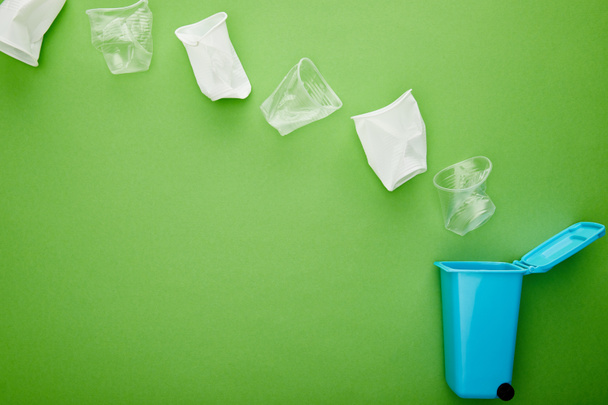 緑の背景に青いごみ箱の近くにくしゃくしゃのプラスチックカップのトップビュー - 写真・画像