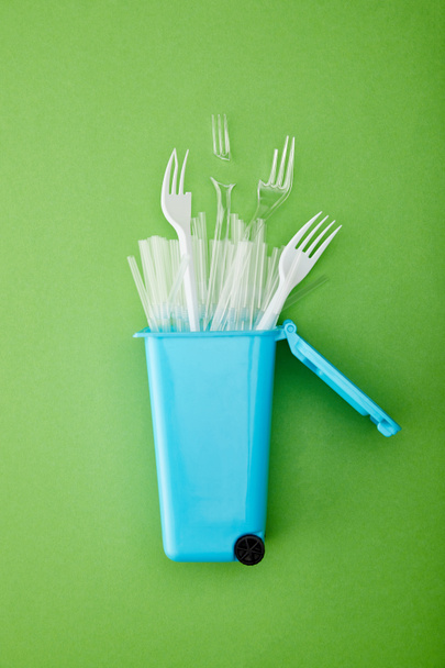 bovenaanzicht van blauwe recycle bin met gebroken plastic vorken en rietjes op groene achtergrond - Foto, afbeelding