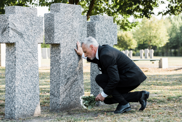 upset senior man in suit sitting near tombstones and holding flowers  - Фото, зображення