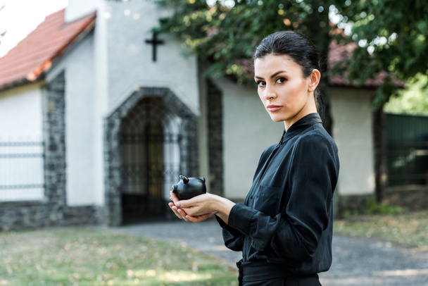 attractive woman holding black piggy bank near church  - Photo, Image