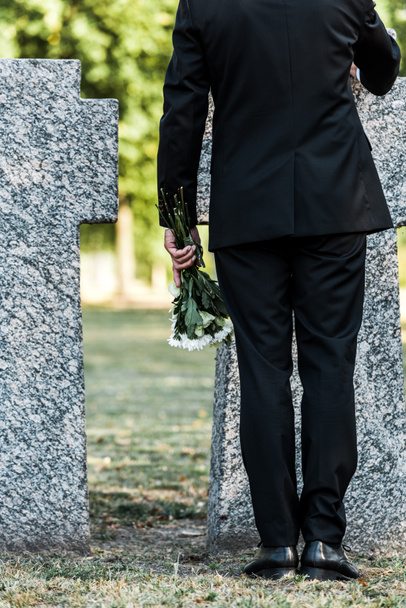 vista recortada del hombre mayor sosteniendo flores y de pie cerca de tumbas
  - Foto, Imagen