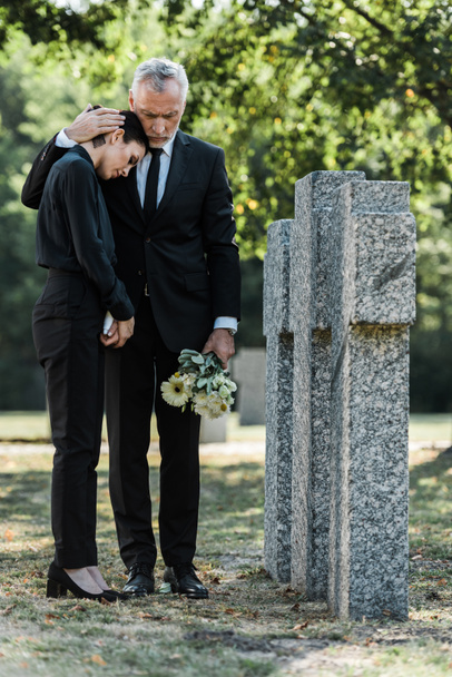senior man holding flowers and hugging upset woman near tombstones - Photo, Image