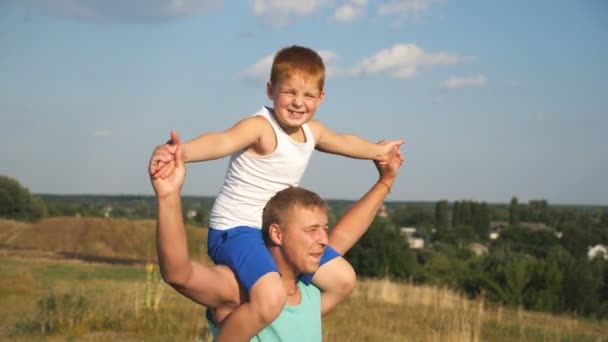 陽気な父親は、笑顔の息子を肩に抱き、晴れた夏の日に牧草地でぐるぐる回る。お父さんと赤毛の子供が芝生で遊んでる。自然の中で過ごす幸せな家族。スローモーション - 映像、動画