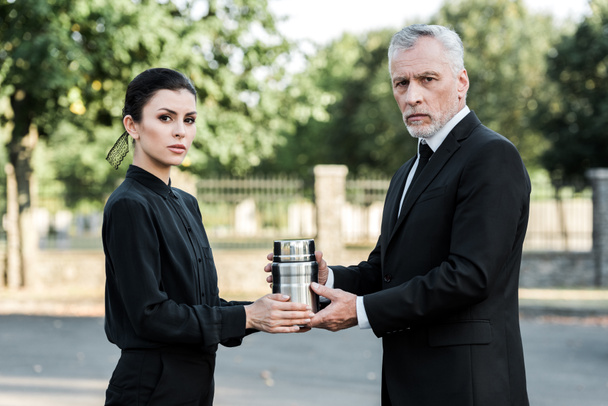frustrado hombre y mujer sosteniendo urna mortuoria y mirando a la cámara
  - Foto, Imagen