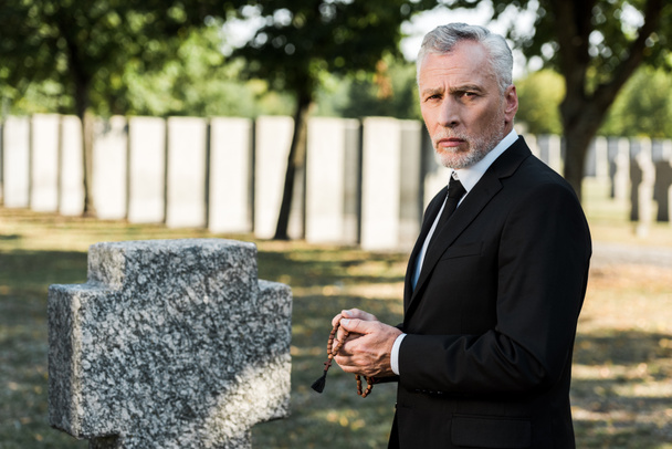грустный мужчина с седыми волосами, держащий четки возле надгробия
   - Фото, изображение