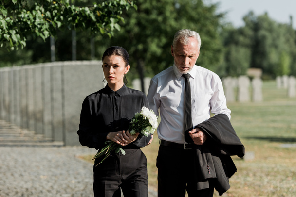 όμορφος ανώτερος άντρας στέκεται με ελκυστική γυναίκα κρατώντας λουλούδια στην κηδεία - Φωτογραφία, εικόνα