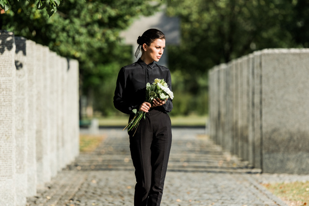 mujer triste sosteniendo flores blancas y caminando en el cementerio
  - Foto, Imagen