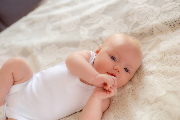 Χαριτωμένο νήπιο μωρό με λευκά ρούχα βρίσκεται σε ένα λευκό κρεβάτι στο σπίτι - Φωτογραφία, εικόνα