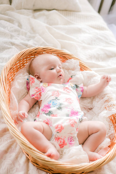 Cute babe baby in floral print clothes lies in a wicker basket on a white bed at home. - Foto, Imagen