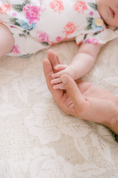 Mom's hands are holding a little cute hand of a newborn daughter at home on a white bed. Happy motherhood - Zdjęcie, obraz