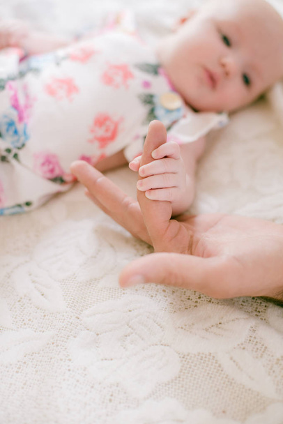 Mom's hands are holding a little cute hand of a newborn daughter at home on a white bed. Happy motherhood - Fotografie, Obrázek