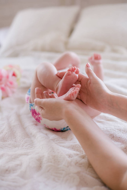 Annemin elleri evde beyaz bir yatakta yeni doğmuş bir kızın küçük sevimli bacaklarını tutuyor. Mutlu annelik - Fotoğraf, Görsel