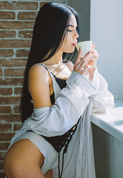 Sensual girl sexy breasts drinking coffee near windowsill. Attractive female lingerie relaxing at home. Passionate mysterious lover. Perfect coffee time. But first coffee. Enjoying morning sunlight - 写真・画像