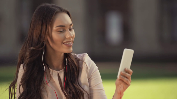 Młoda kobieta siedzi na trawie i słuchania muzyki na smartfonie - Materiał filmowy, wideo