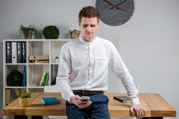 Attractive young businessman using a cell phone. Cheerful Businessman With Smartphone Standing In An Office. Portrait of man in office with telephone. Businessman using phone at workplace in office - Photo, image