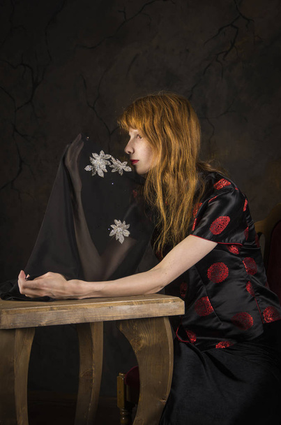 Red-haired girl in dark clothes sitting at the table. - Фото, зображення