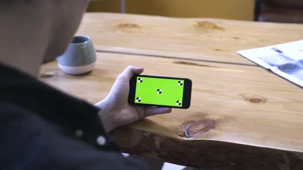 Pohled z ramene mladého muže v černé košili, sedící u dřevěného stolu, a pohled na zelenou obrazovku, která je Chroma klíčem. Skladní záběry. Nový koncept technologie smartphone - Záběry, video