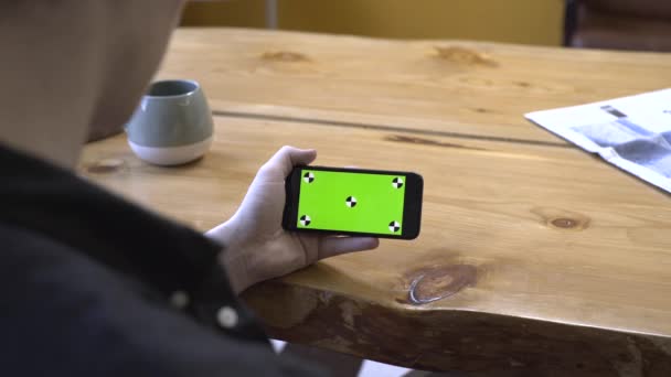 Pohled z ramene mladého muže v černé košili, sedící u dřevěného stolu, a pohled na zelenou obrazovku, která je Chroma klíčem. Skladní záběry. Nový koncept technologie smartphone - Záběry, video