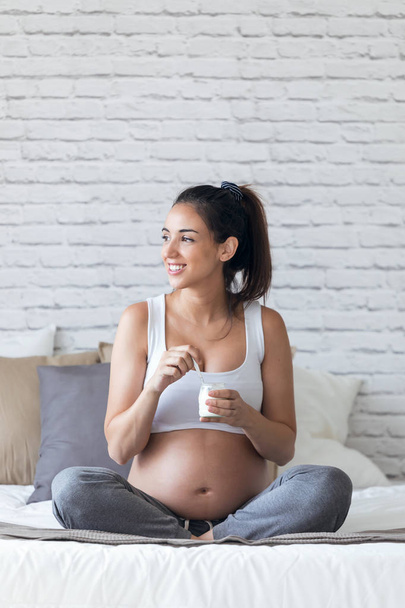 Kaunis nuori raskaana oleva nainen syö jogurttia istuessaan sängyssä kotona
. - Valokuva, kuva