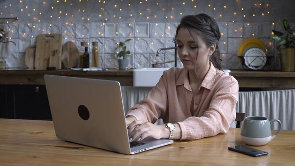 Szép fiatal nő ül a konyhában, fából készült asztal és gépelés rajta laptop a csésze teát vagy kávét. Stock Footage. Modern berendezésű otthon belső tér a háttérben - Fotó, kép