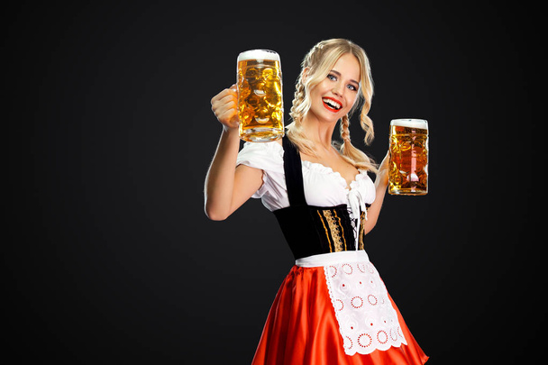Happy sexy oktoberfest girl waitress, wearing a traditional Bavarian or german dirndl, serving two big beer mugs with drink isolated on black background. - Фото, изображение