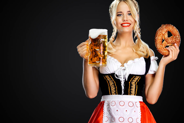 Улыбающаяся молодая сексуальная Октоберфест официантка, одетая в традиционное баварское или немецкое платье, подающая большие кружки пива с напитком и бретцелем, изолированные на черном фоне
. - Фото, изображение