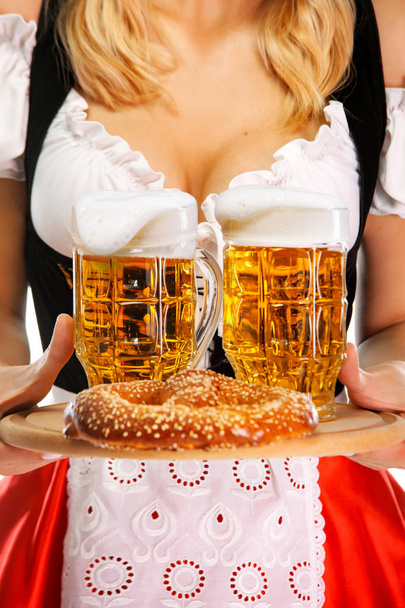 Closeup of young sexy Oktoberfest girl waitress, wearing a traditional Bavarian or german dirndl, serving two big beer mugs with drink and bretzel. - Fotografie, Obrázek