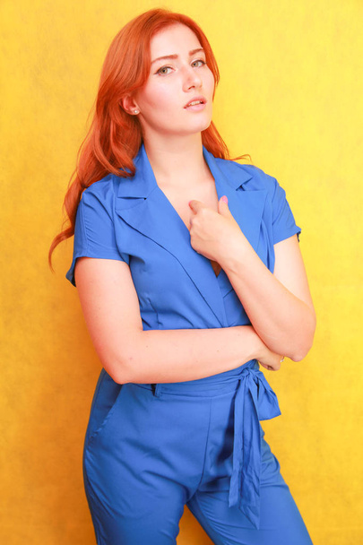 pretty red hair woman in blue fashion overall standing on yellow background - Zdjęcie, obraz