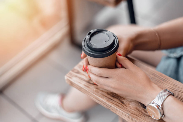 Paper coffee cup to go in womans hands with red manicure while sitting in cafe. - Foto, Bild