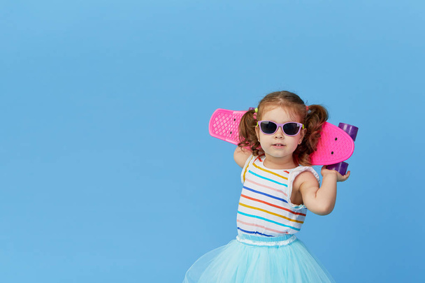 Cool little toddler girl in bright clothes and sunglasses carrying a skateboard over his shoulder and looking at the camera isolated on blue background - Фото, изображение