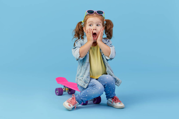 Cool little toddler girl in denim clothes sits on the skateboard and funny looking at the camera isolated on blue background - Φωτογραφία, εικόνα