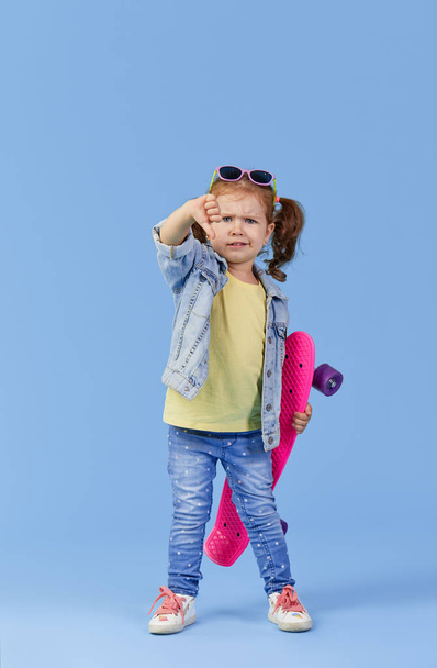 Stylish cool serious little girl child with skateboard over blue background.  showing thumb down displeased. Space for text - Photo, image