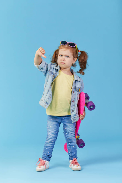Stylish cool serious little girl child with skateboard over blue background.  showing thumb down displeased. Space for text - Foto, Imagem