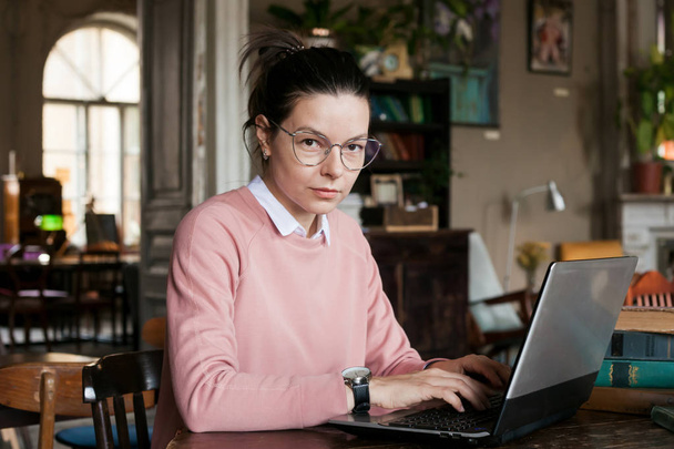 Voorbereiding van het examen. Aantrekkelijke brunette student meisje in glazen en een roze trui, met een lichte glimlach zit op een laptop. - Foto, afbeelding