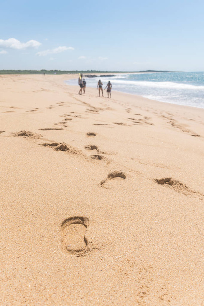 Fußabdrücke entlang der Sandküste, eine Gruppe von vier Personen hängt am Strand, der Ozean hat nur eine einzige Welle, am Horizont stehen einige Pflanzen, der Himmel hat nur wenige Wolken - Foto, Bild