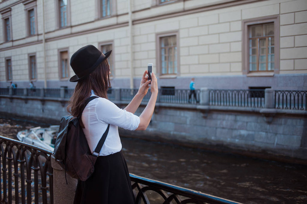 Tourist in the city takes a photo on smartphone. A young woman in a black hat and a white shirt, - Fotografie, Obrázek