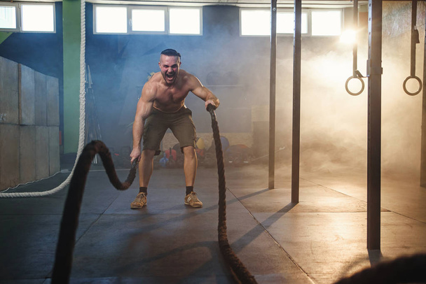 Handsome strong man doing exercises for the muscles with battle ropes in fitness gym. - Foto, afbeelding