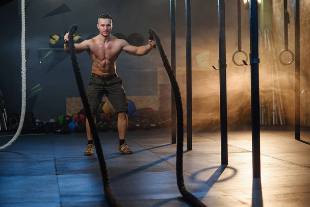 Красивый сильный мужчина делает упражнения для мышц с боевыми веревками в фитнес-зале
. - Фото, изображение