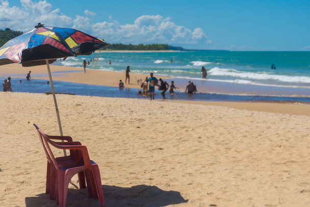 Una silla roja de plástico bajo una sombrilla con patrones en una playa concurrida de Trancoso, Porto Seguro, Bahía, con turistas nadando en el encuentro de las aguas de manglar con el mar
 - Foto, imagen