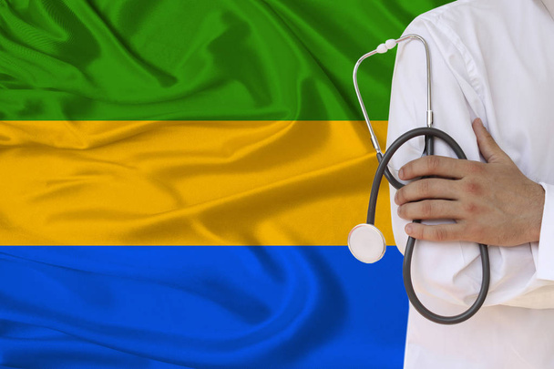 partial photograph of a doctor in uniform against the background of the national flag of the state of Gabon on delicate shiny silk, the concept of medical care and insurance in the country, copy space - Photo, image