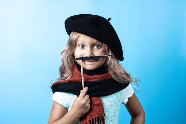 Funny girl in black beret, scarf and mustache on a stick on blue background - Valokuva, kuva