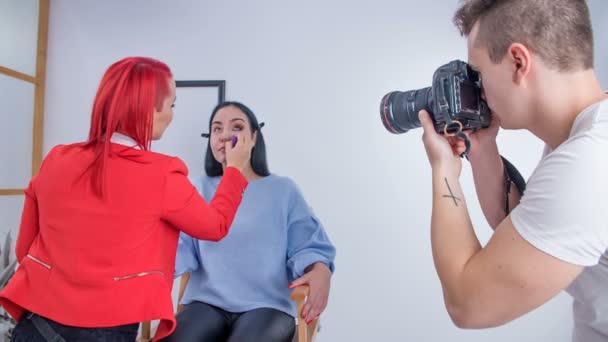Een make-up kunstenaar brengt vloeibaar poeder aan op het gezicht van haar cliënt, vooral in de gebieden rond haar ogen, neus en wangen. Een fotograaf maakt foto 's tijdens een make-up sessie. - Video
