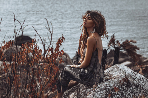 beautiful young boho style woman sitting on stone at sea  - 写真・画像