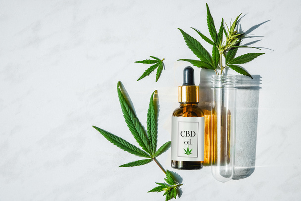 Glass bottle with cannabis oil with label and a test tube with hemp leaves on a marble background. Copy space. - Fotografie, Obrázek
