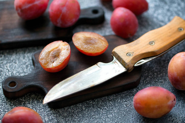 Whole and sliced ripe plums, a knife with a wooden handle on a wooden board on a grey table.. Preparation of the pie. Front view - Fotó, kép