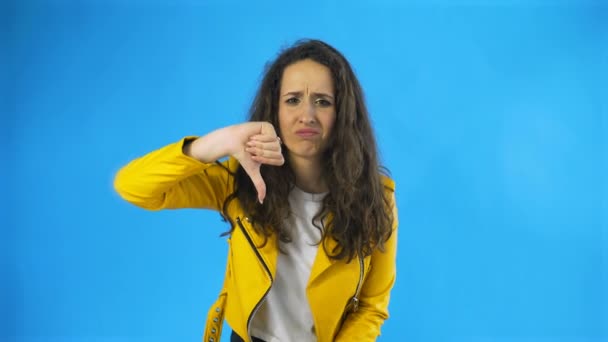 Retrato de mulher séria em casaco amarelo mostrando polegares para baixo sinal de não gostar no Estúdio com fundo azul
. - Filmagem, Vídeo