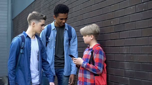 Senior pupils threatening younger boy with phone, bullying nerd in backyard - Foto, imagen